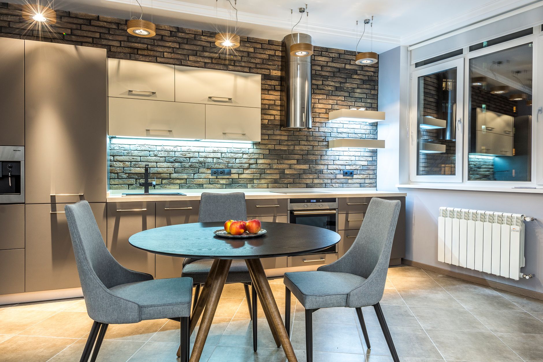 Cuisine moderne avec table ronde bleue, chaises grises et îlot blanc face à un mur de briques