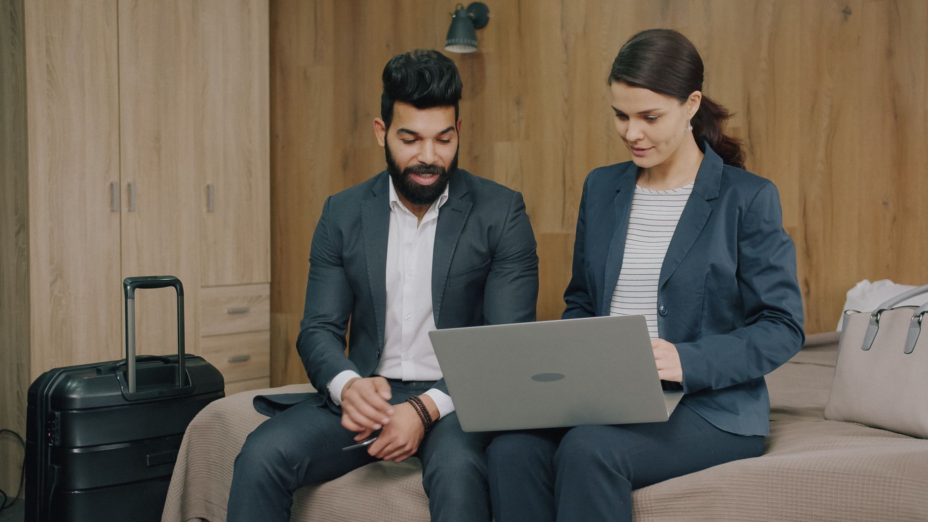 Two business professionals in dark blazers reviewing a laptop together while seated on a bed in a hotel room with luggage nearby
