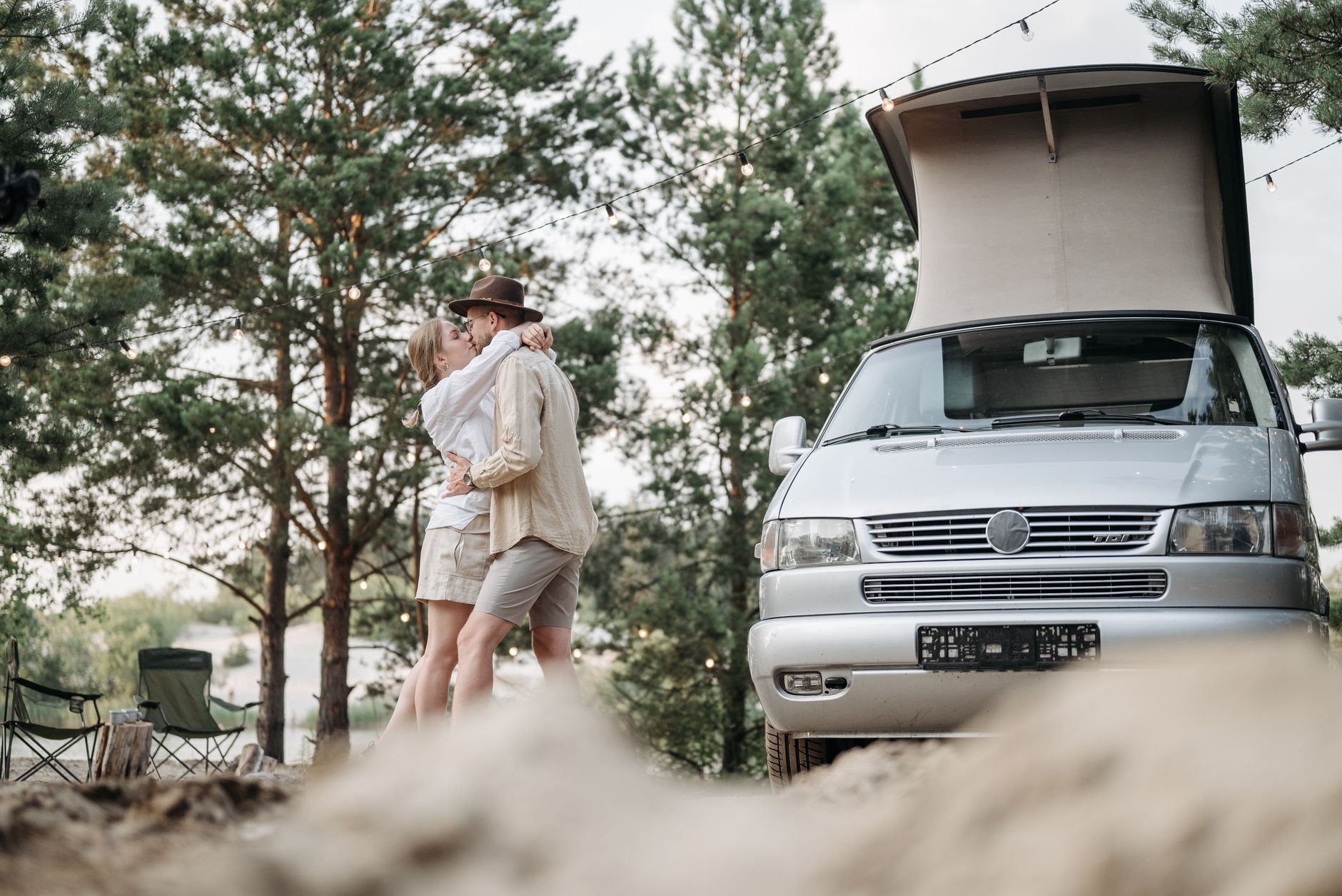 Couple embracing outdoors next to a silver campervan with pop-up roof in a forested campsite setting