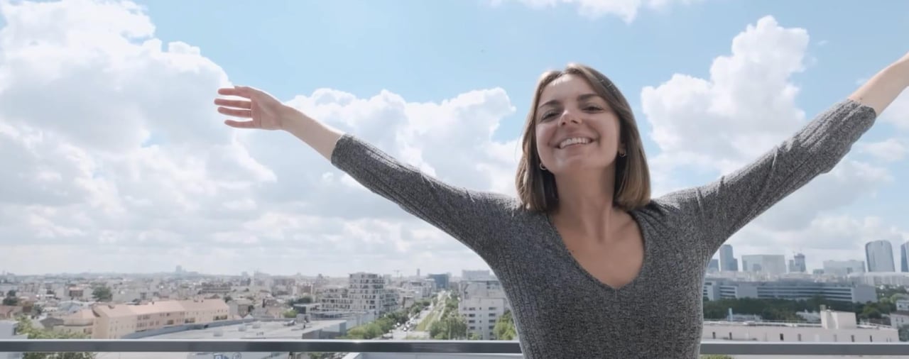 Femme souriante les bras levés face à la ville depuis un balcon par temps ensoleillé