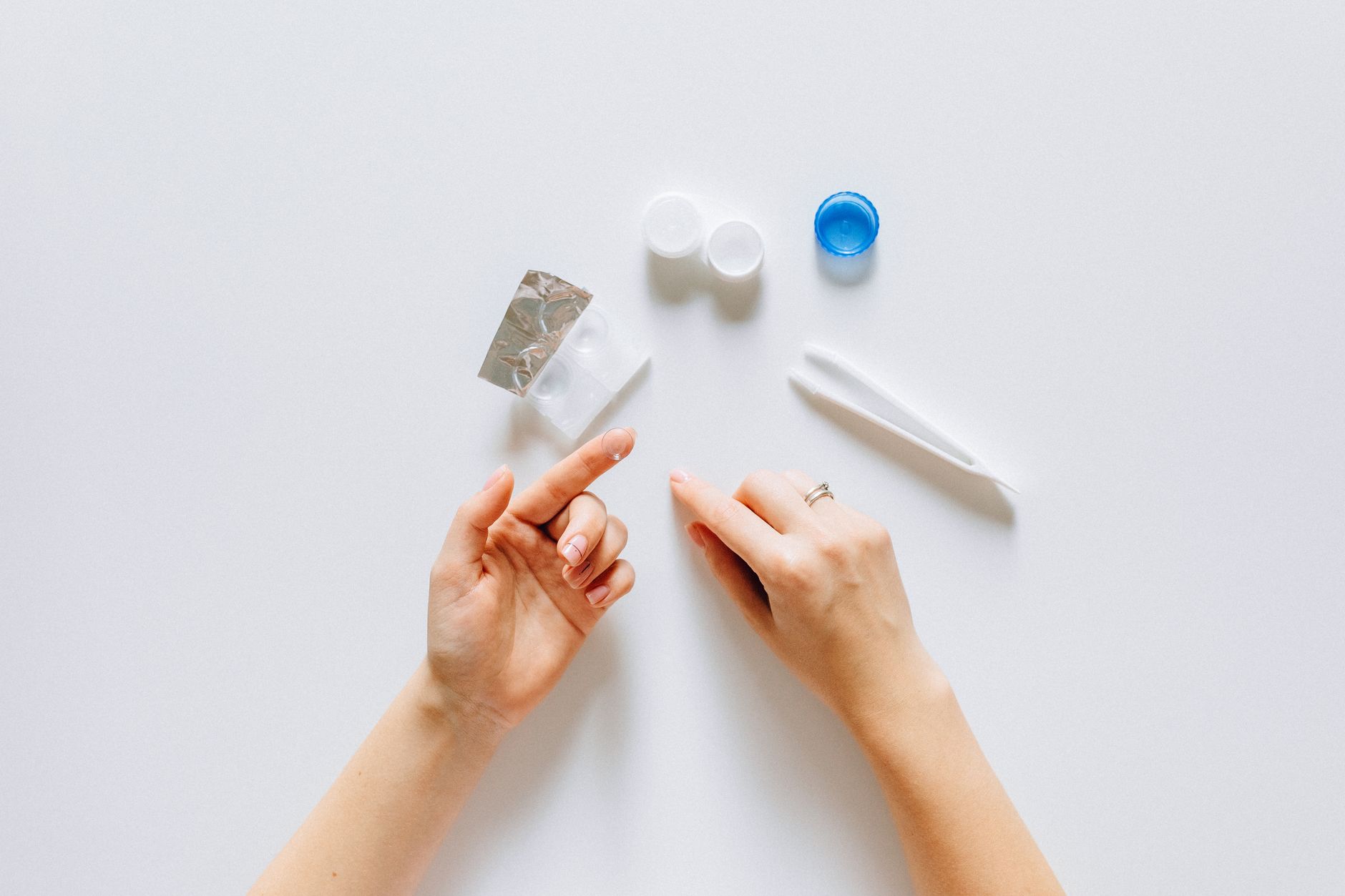 Overhead view of hands with contact lens supplies including white bottles, a foil packet, tweezers, and a blue solution container on white surface
