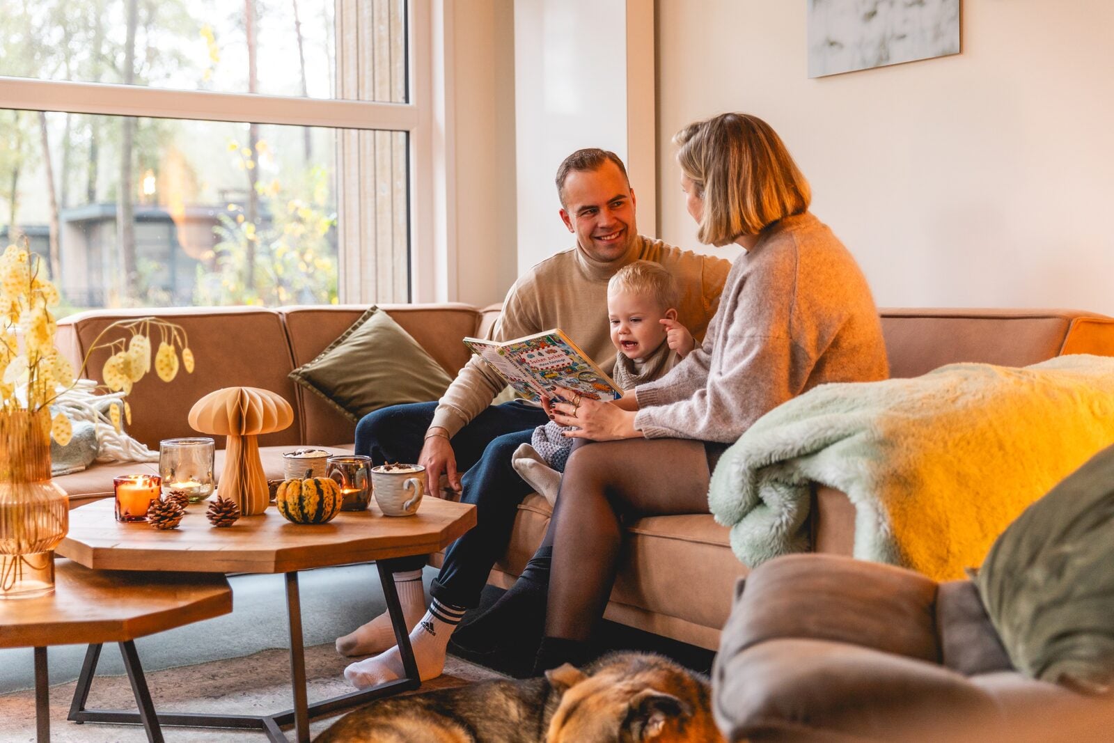 Familie mit Kleinkind liest gemeinsam auf braunem Sofa in gemütlichem Wohnzimmer mit Herbstdekoration