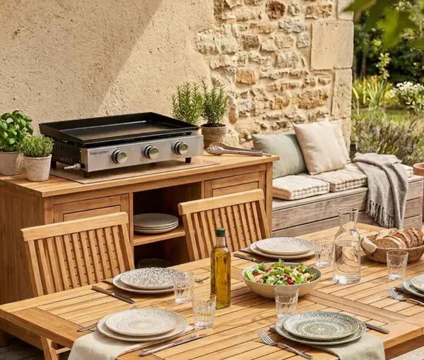 Cuisine d'extérieur en bois avec barbecue et table de repas dressée face à un mur en pierre