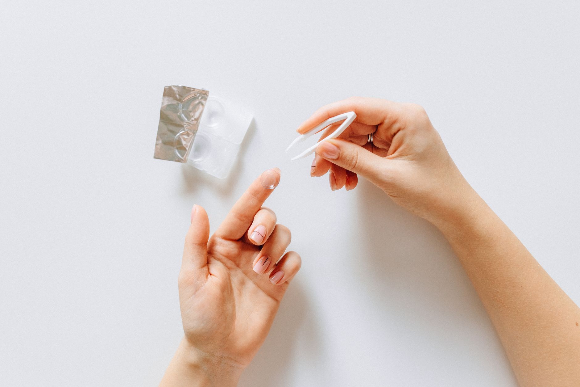 Close-up of hands holding tweezers near contact lens supplies with a foil packet and white bottle visible on white background