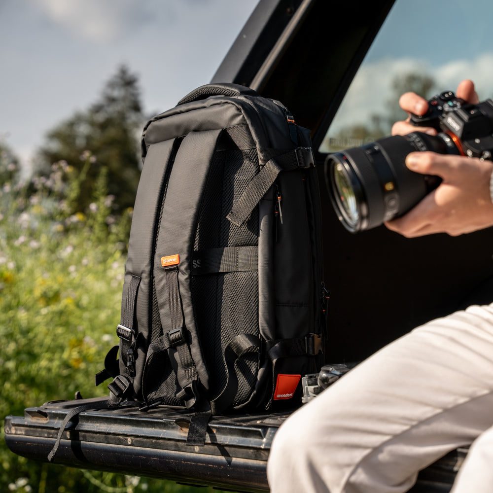 Black travel backpack positioned on car roof rack next to person holding professional camera lens