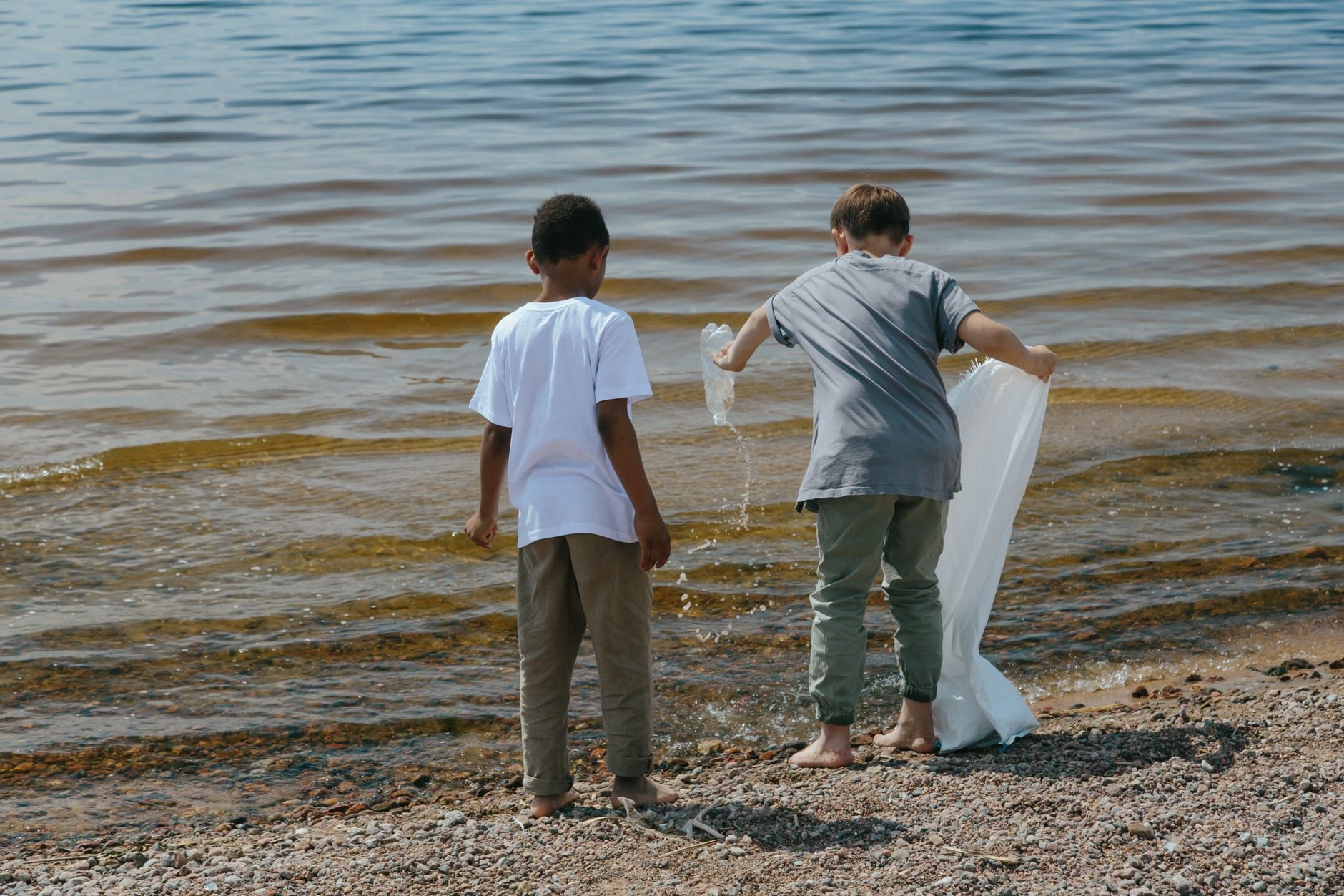 Two children collecting plastic waste in white bags on a beach near the water