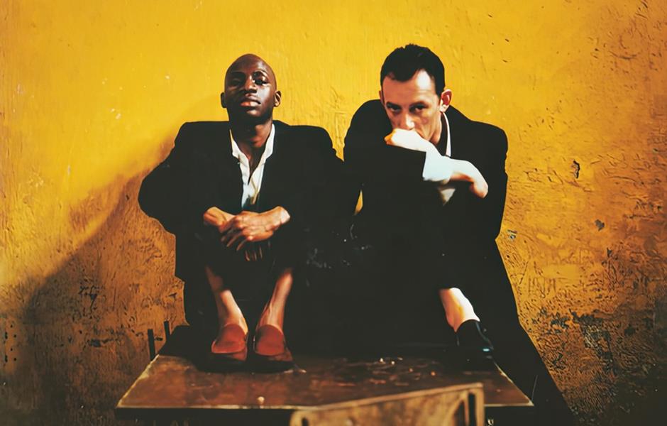 Deux hommes en costumes noirs assis contre un mur jaune ocre