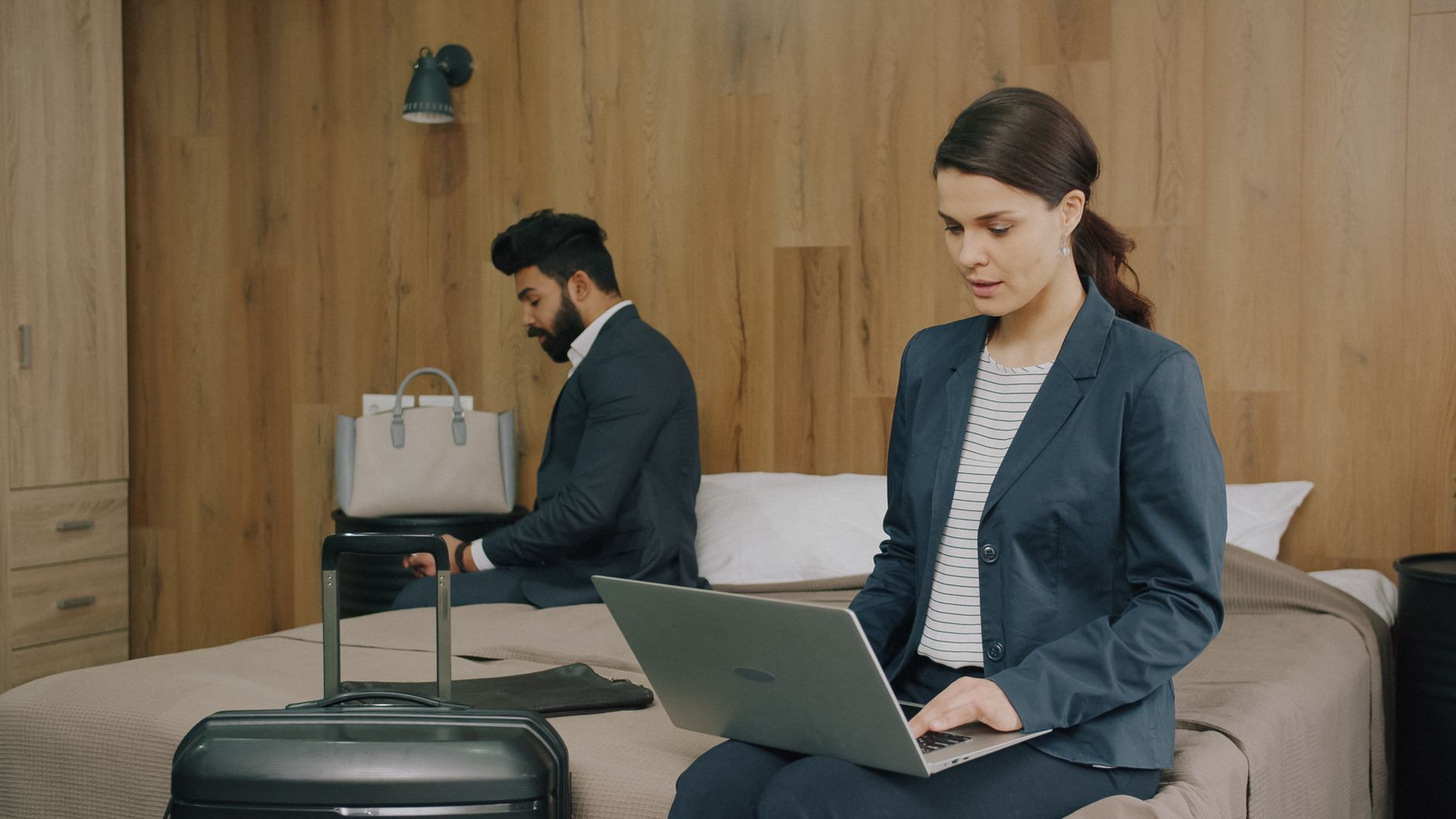 Woman in business attire working on a laptop while seated on a bed, with a man in the background handling luggage in a hotel room
