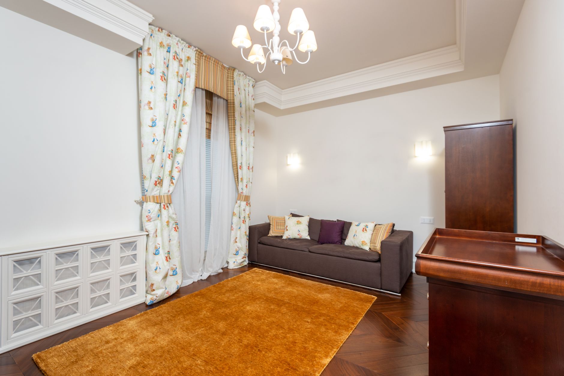Elegant living room with dark brown sofa, white geometric cabinet, patterned curtains, wooden rug, and chandelier overhead