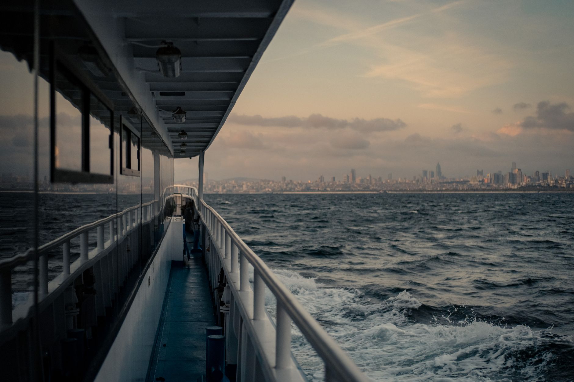 Aussicht von der Reling einer Fähre auf das Meer und eine Stadtskyline am Horizont bei Sonnenuntergang.