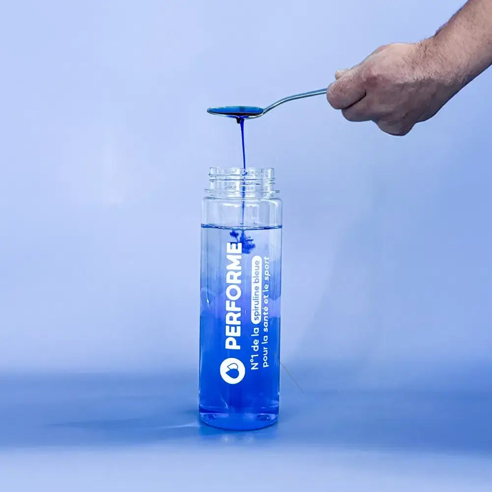 Main contenant une cuillère de spiruline bleue au-dessus d'une bouteille avec étiquette blanche et bleue sur fond bleu