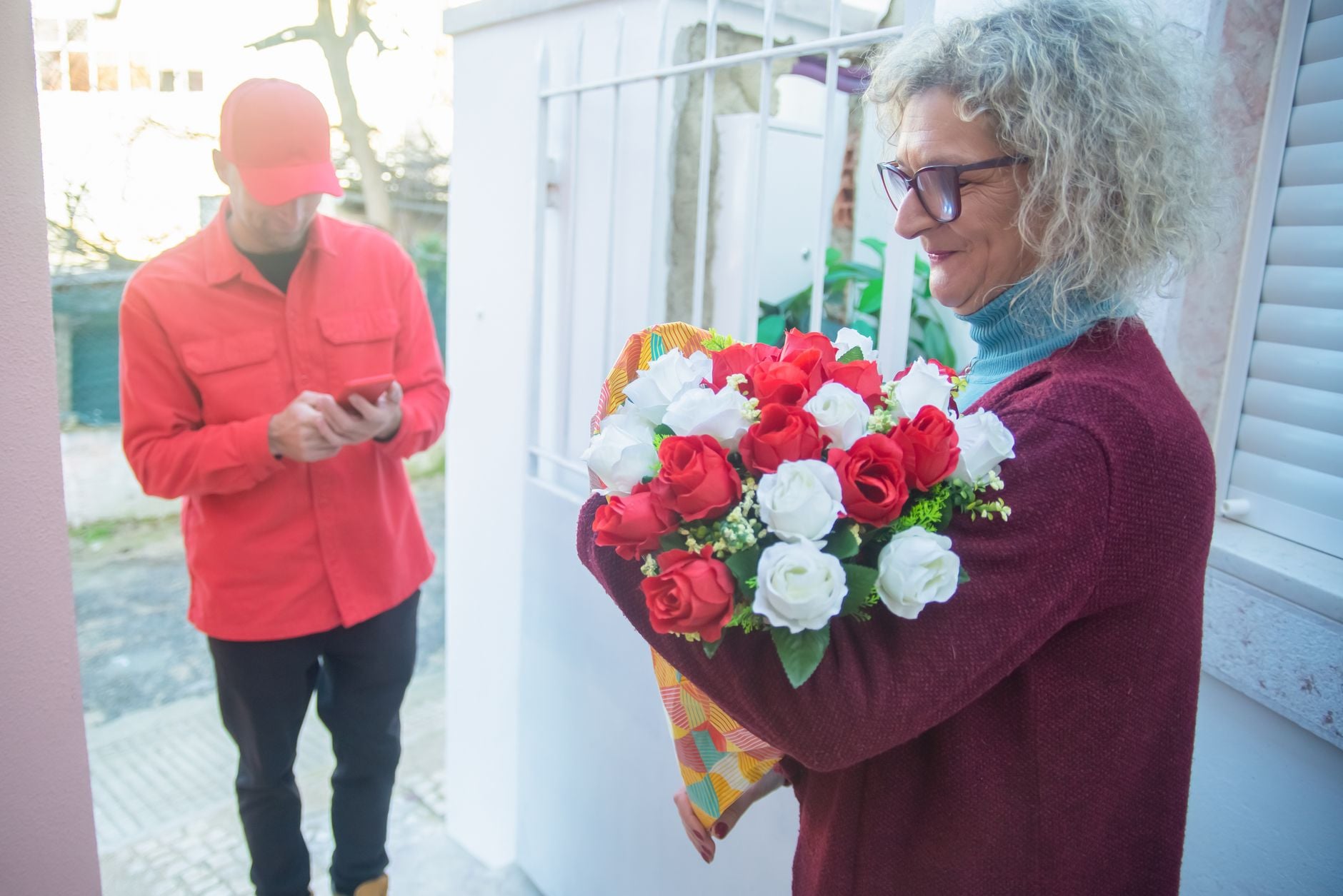 Lieferbote in rotem Hemd überreicht Frau roten und weißen Blumenstrauß an der Haustür.